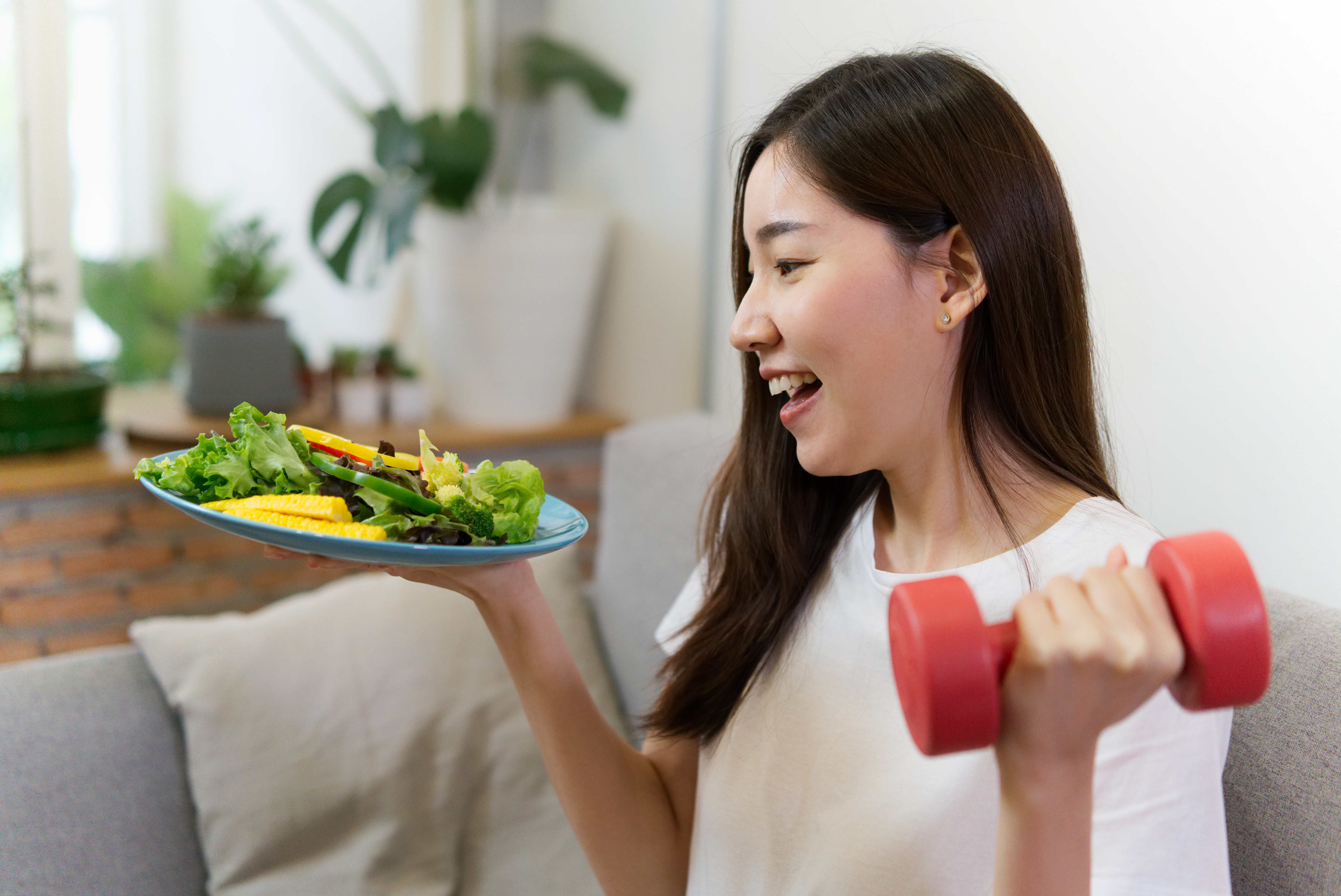 Young Asian girl holding salad and red dumbbell is sitting on sofa with smile face. Woman healthy concept.