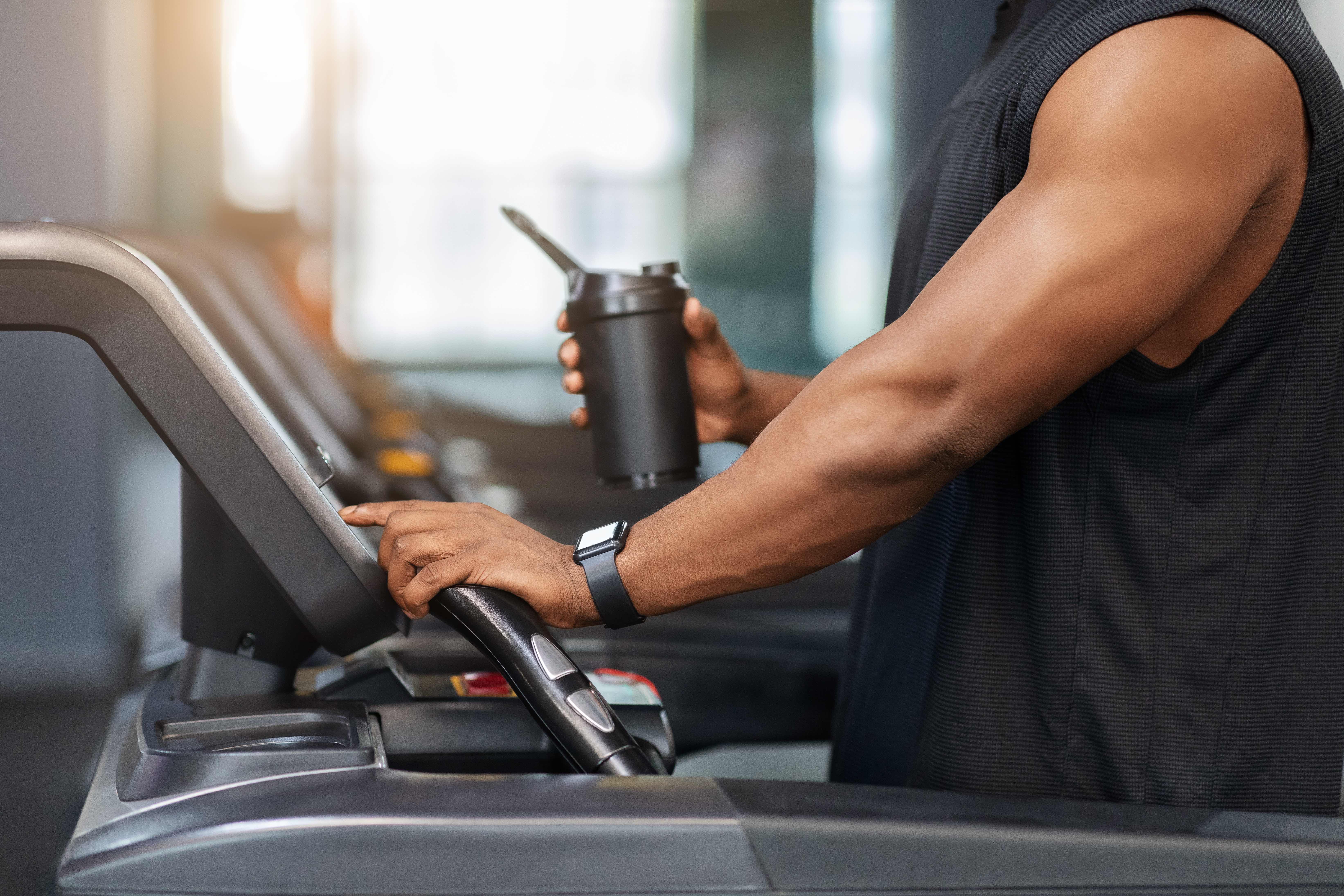 Cropped of muscular african american man holding sports drink, standing on treadmill, drinking protein before running, copy space. Unrecognizable black guy bodybuilder jogging at gym