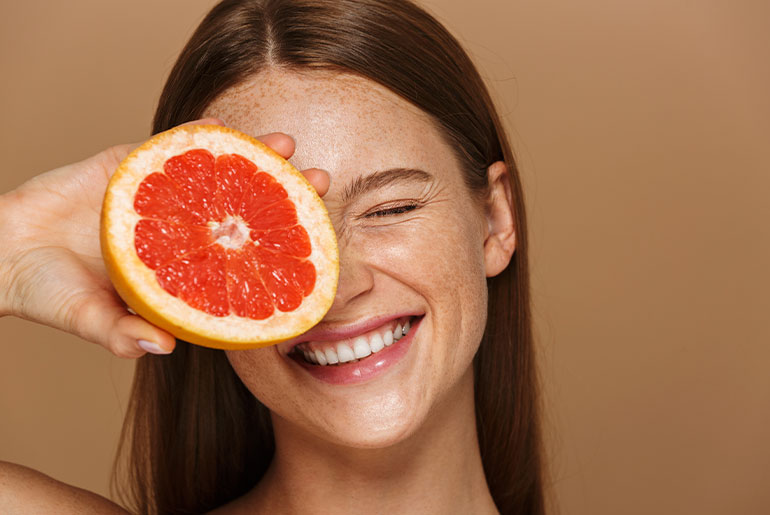 A portrait shot of a woman holding a half of a grapefruit in front of her left eye and smiling